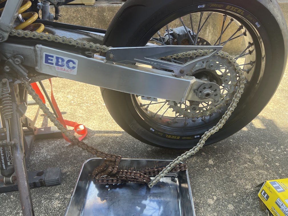 バイクのリアスプロケットに掛けた新品のチェーンと、トレーに下ろした赤サビのある古いチェーン